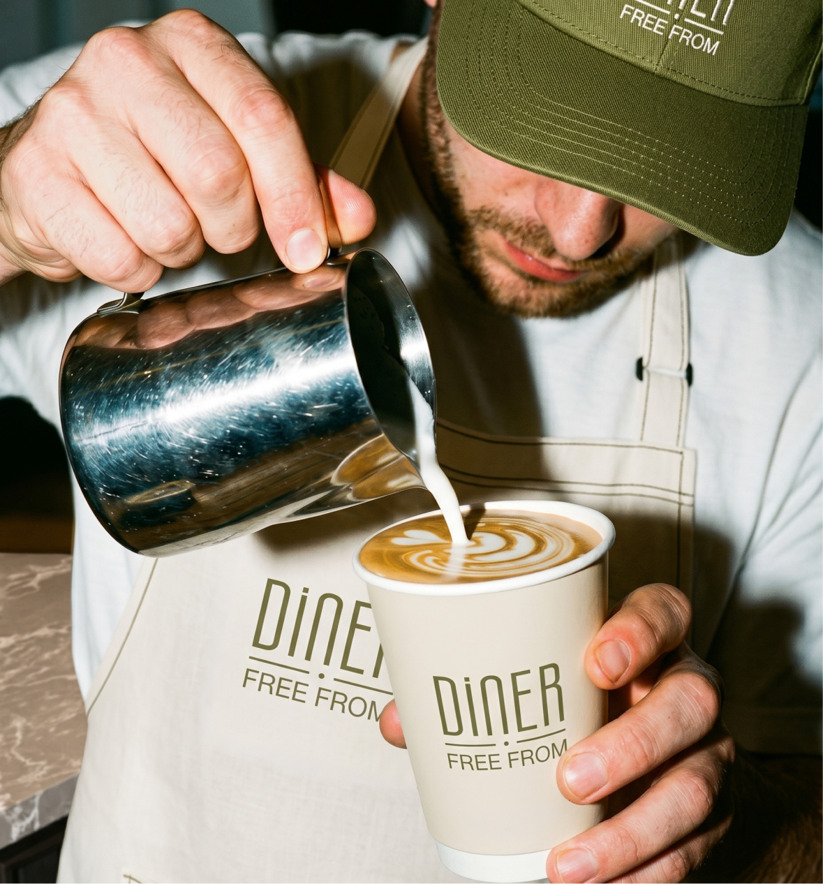Barista in a Diner Free From cap and apron pouring latte art into a branded cup