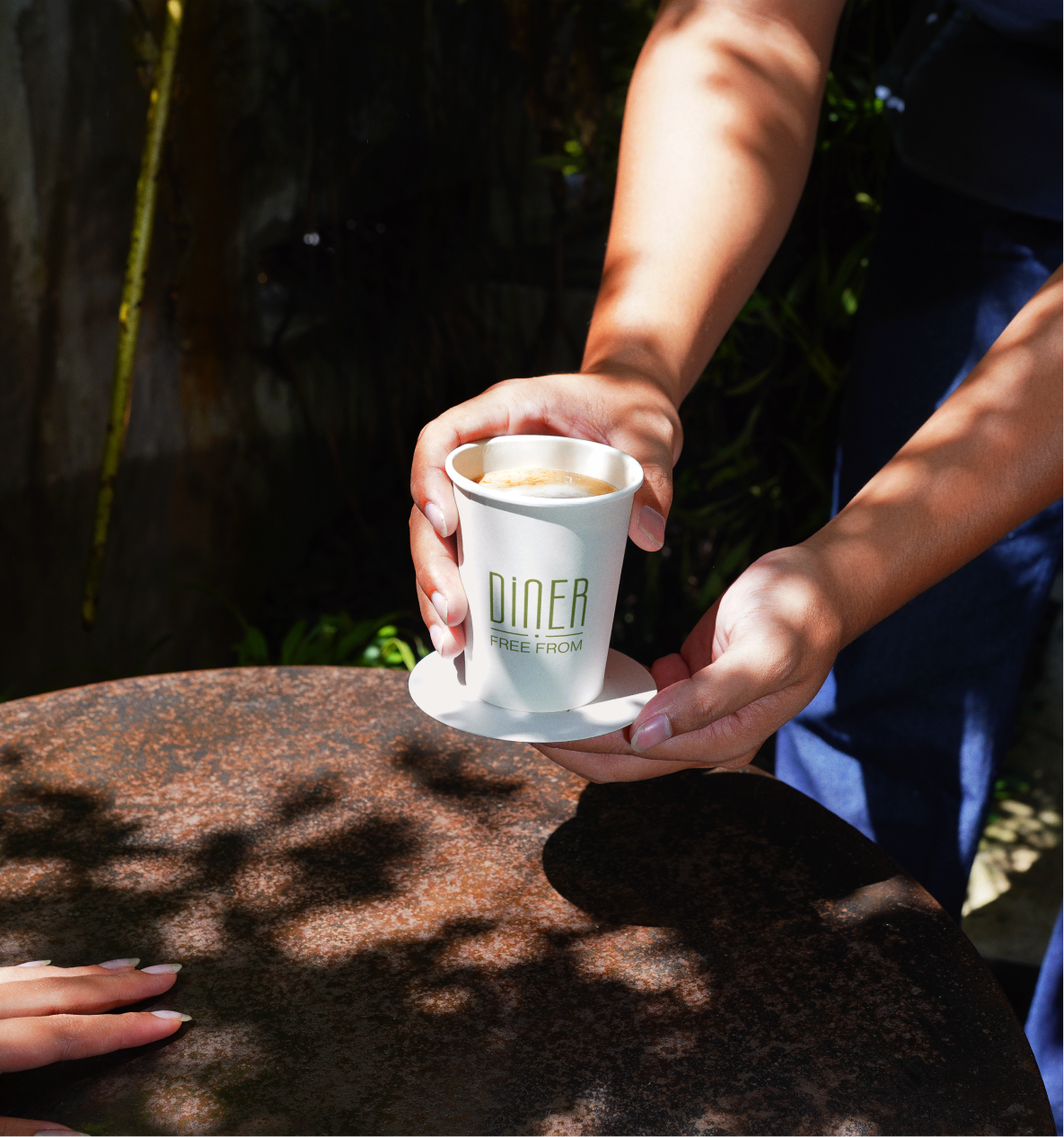 Hands passing a Diner Free From branded coffee cup over an outdoor table
