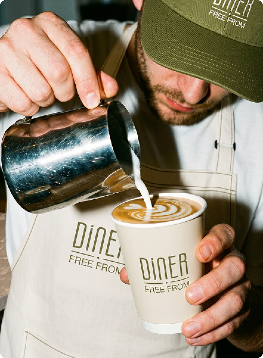 Mask group DFF Barista in a Diner Free From branded cap and apron pouring latte art into a takeaway cup