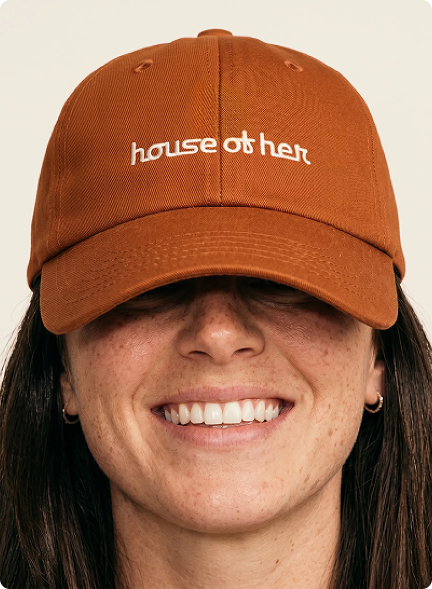 Mask group HOF Close-up of a woman smiling in a burnt-orange baseball cap embroidered with the House of Her logo