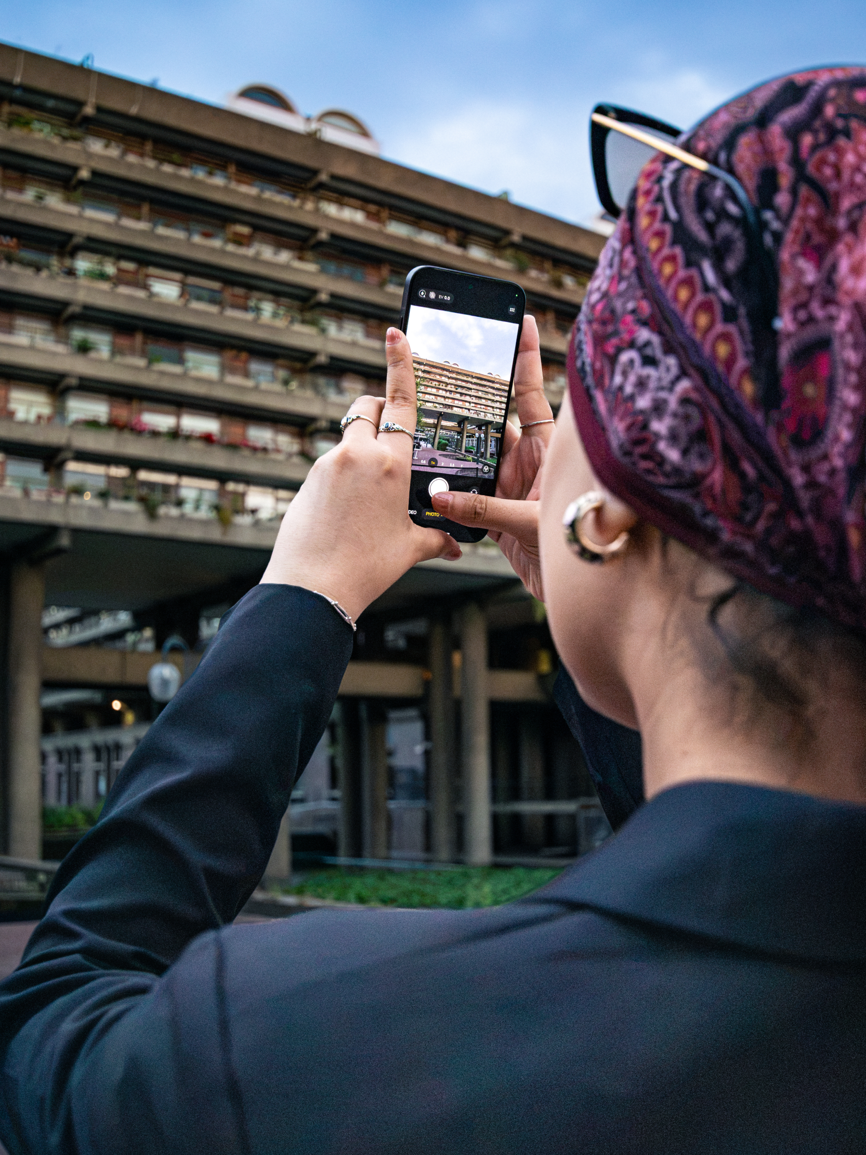Low-angle photograph of a woman holding a phone against her chest, Barbican Centre behind her