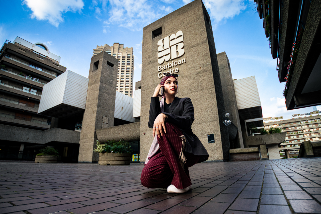 Woman on a phone call leaning against a curved bench at dusk, Barbican architecture behind her