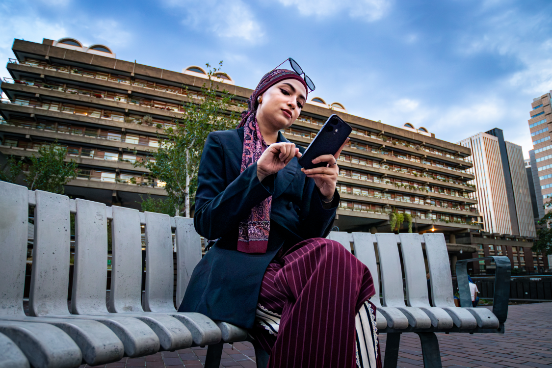 Woman seated on a bench looking at her phone, Barbican residential block behind her