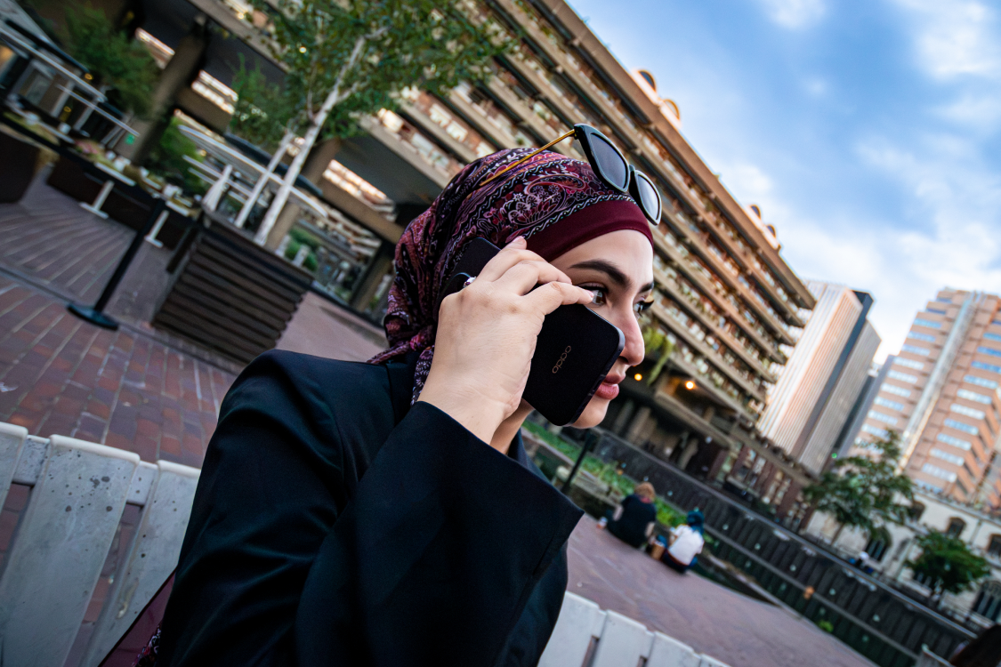 Close-up profile of a woman holding a phone to her ear, Barbican terrace behind her